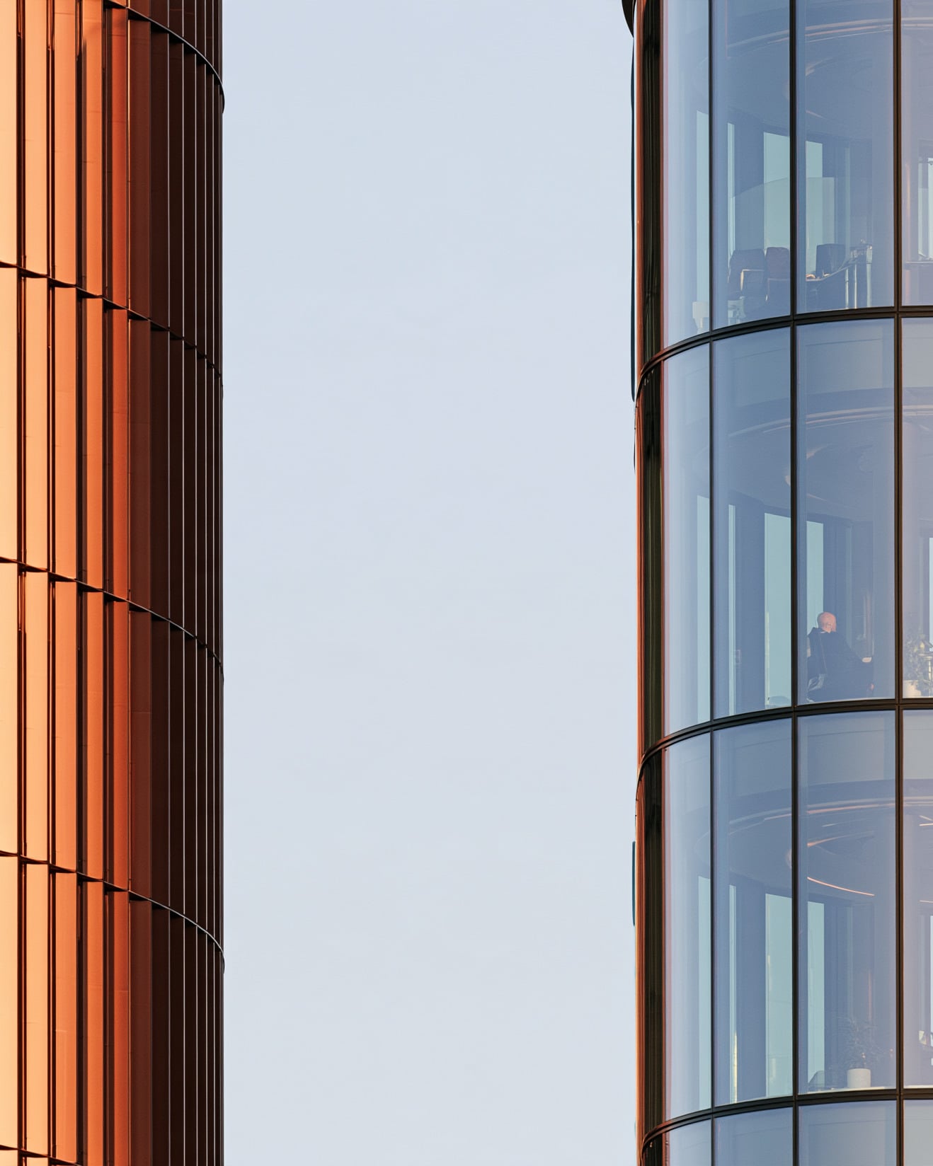A copper-clad tower beside a glass office tower, people at work behind the glass